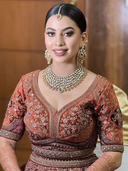 A smiling portrait of a bride in a rich, rust-colored lehenga. The makeup is warm and glamorous, with a focus on defined eyes.