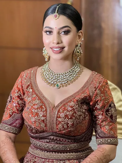 A smiling portrait of a bride in a rich, rust-colored lehenga. The makeup is warm and glamorous, with a focus on defined eyes.
