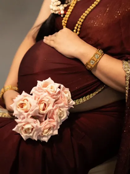 A close-up, artistic shot celebrating traditional Indian maternity style. This image highlights the beauty of a rich maroon saree, intricate gold jewelry, and the gentle embrace of the baby bump.