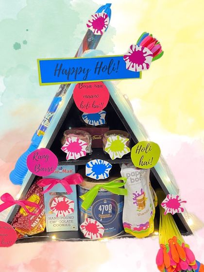 A stylish triangular hamper for Holi. This black A-frame stand is filled with cookies, popcorn, and other goodies, and decorated with pichkaris and colourful accents.