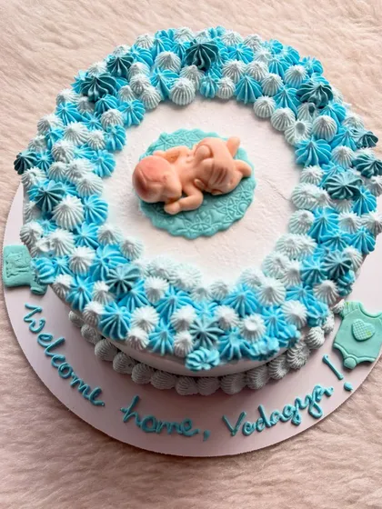 A sweet welcome cake for a newborn baby, with a delicate blue and white piped wreath and a tiny sleeping baby in the center.