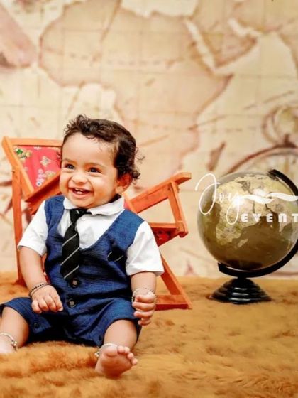 A charming traveler-themed photoshoot for a one-year-old. Dressed in a smart vest and tie, the baby sits in front of a world map backdrop with a globe and vintage props, ready for an adventure.
