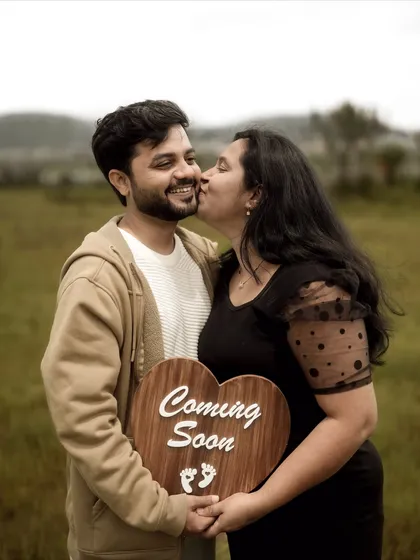 A joyful and candid moment. The mom-to-be gives a happy kiss on the cheek while they both hold a heart-shaped "Coming Soon" sign, perfectly capturing the excitement of this new chapter.