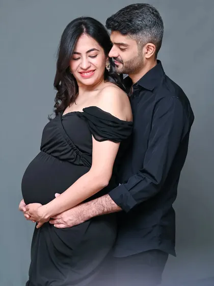 An elegant studio portrait of a couple dressed in black. The dad-to-be embraces his partner from behind, creating a feeling of love and protection.