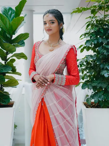 This vibrant half-saree combines a bright orange skirt with a sheer, light pink davani. The look is completed with a custom red blouse featuring intricate silver embroidery on the full sleeves.