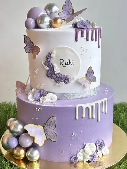 This elegant two-tiered birthday cake combines soft violet and white whipped cream frosting. We decorated it with delicate butterflies, chocolate drips, and shimmering spheres for a sophisticated and beautiful celebration cake.