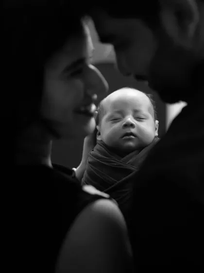 An intimate black and white close-up of new parents gazing lovingly at their sleeping newborn held between them.
