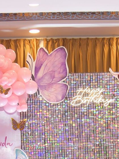A close-up of a butterfly-themed birthday backdrop. The iridescent shimmer wall is decorated with large butterfly cutouts and a white neon "Happy Birthday" sign, creating a sparkling effect.