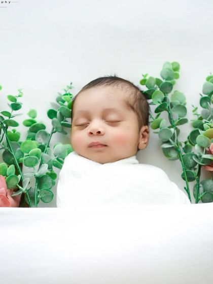 A touch of greenery adds a fresh, natural feel to this minimalist setup. The baby is wrapped in white, lying on a clean white surface.