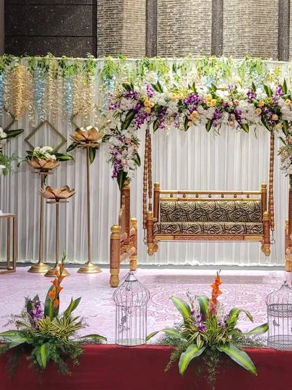 Another angle of the traditional swing (jhoola) stage, highlighting the intricate floral work and the golden lotus lamp stands. The hanging floral curtains in the background add a delicate touch.