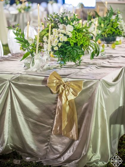 A detail of a tablescape from a Goa dinner party. The table is draped in a beautiful silver-grey fabric with a large gold bow, and topped with a fresh green and white floral arrangement.