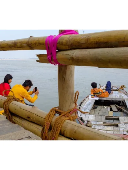 A scene from Varanasi where a couple takes a selfie by the Ganga, while another person relaxes in a boat. The image captures the blend of modern life and traditional relaxation on the ghats.