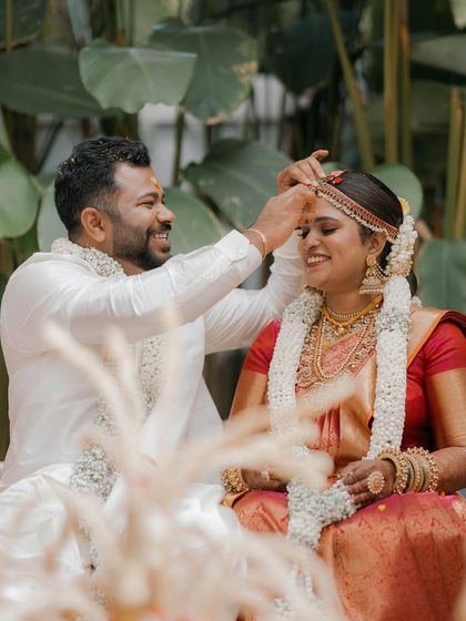 A candid moment from a morning wedding ceremony. The groom lovingly places the sindoor on the bride, with the lush green foliage providing a serene and natural background.