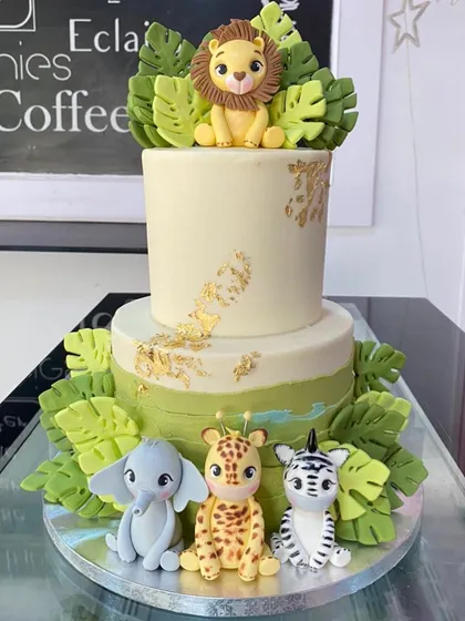 A sophisticated two-tiered safari cake with gold leaf accents. This design features cute, stylized fondant animals including a lion, elephant, giraffe, and zebra.