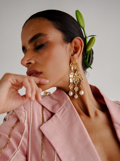 A close-up beauty shot featuring large, floral-inspired pearl drop earrings. The styling is romantic, with a fresh lily tucked into the model's hair to complement the theme.