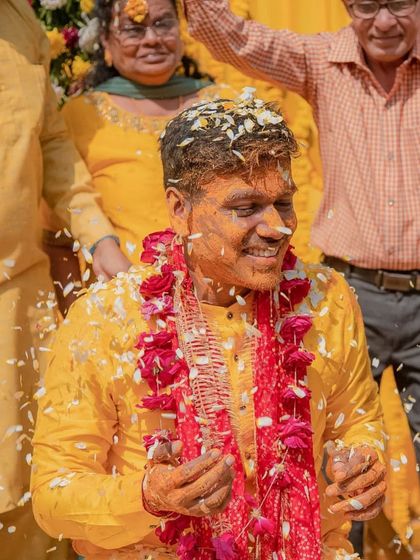A portrait of the groom smiling, covered in turmeric and flower petals, with a garland of red flowers.