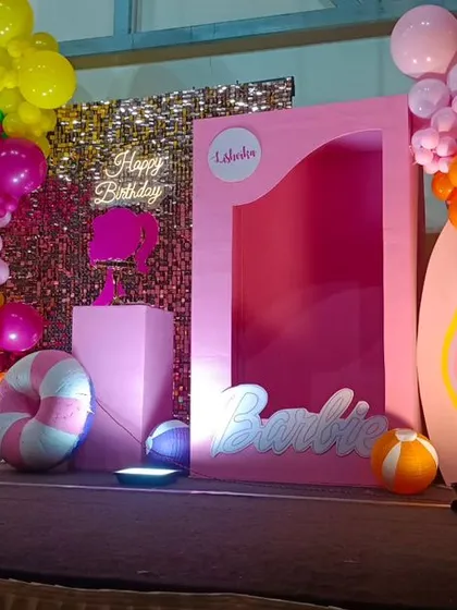 A vibrant, tropical Barbie-themed birthday party. This setup features a pink "doll box" photo booth, a sequin backdrop, and colorful balloon clusters with palm tree cutouts.