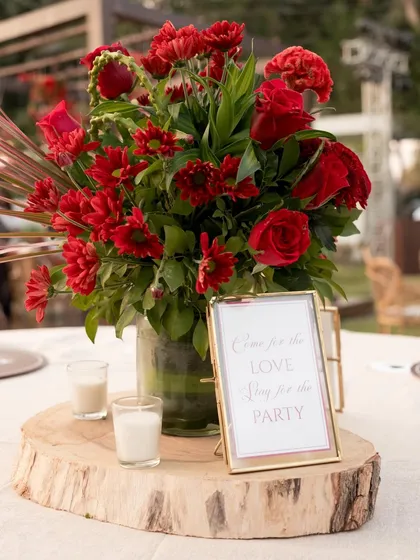 A rustic yet elegant centerpiece featuring a log slice, a vibrant red floral arrangement, and a simple, sweet sign.