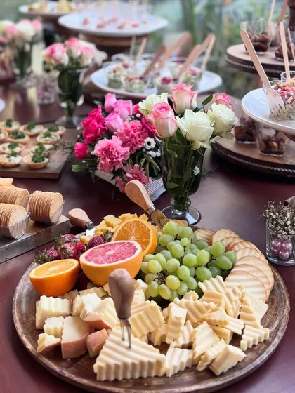 A close-up of a cheese platter from a high-tea event, featuring ridged cheeses, fresh grapes, strawberries, and grapefruit. The arrangement is complemented by beautiful pink and white floral bouquets.