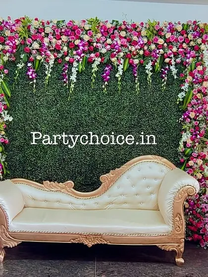 A beautiful floral photo wall with a green mat base and a border of pink and purple flowers. The chaise lounge provides elegant seating for a puberty ceremony or half saree function.