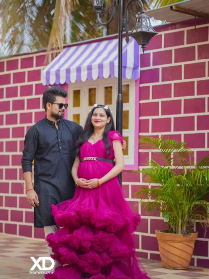 A stylish couple posing against a colorful pink brick wall. The tiered magenta gown and coordinated outfits create a chic and modern urban vibe.