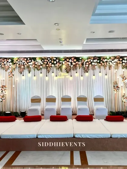 An all-white wedding stage featuring traditional floor seating, a canopy of white and blush florals, and hanging light bulbs for a serene and elegant atmosphere.