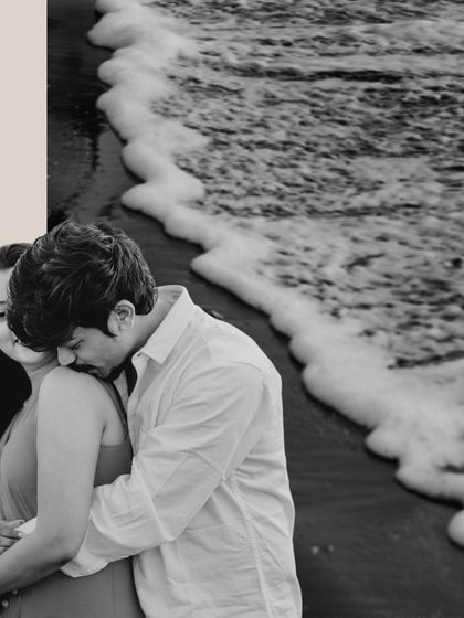 A tender black and white photo of the groom embracing the bride from behind, with the texture of the sea foam in the background.