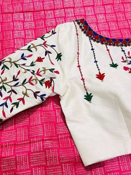 This elegant white blouse features colorful bird and floral motifs embroidered with threadwork. The neckline and sleeves are highlighted with a traditional red and blue border, perfect for pairing with a classic Kanchipuram saree.