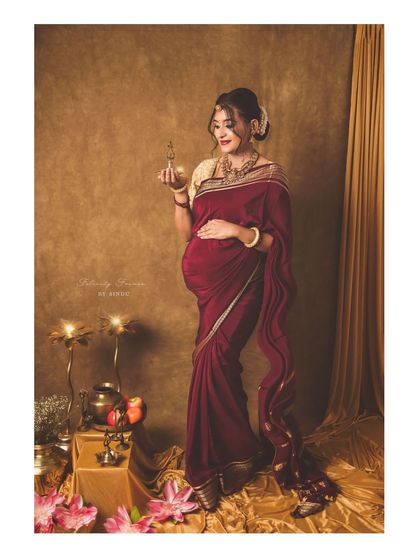 A full-length portrait of a mother-to-be in a rich maroon saree, holding a traditional lamp. The setup, complete with brass vessels and a golden backdrop, evokes a sense of timeless elegance.