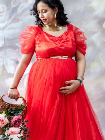 A lovely studio portrait against a floral painted backdrop. The mom-to-be looks elegant in a red tulle gown, holding a basket of fresh flowers.