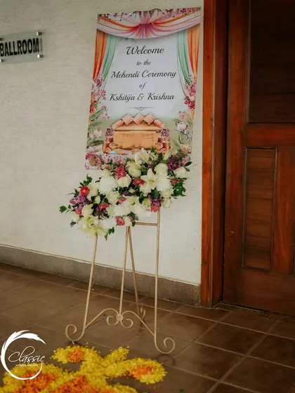 A simple and elegant welcome sign for the Mehendi ceremony, placed on an easel and adorned with a fresh floral arrangement to greet guests.