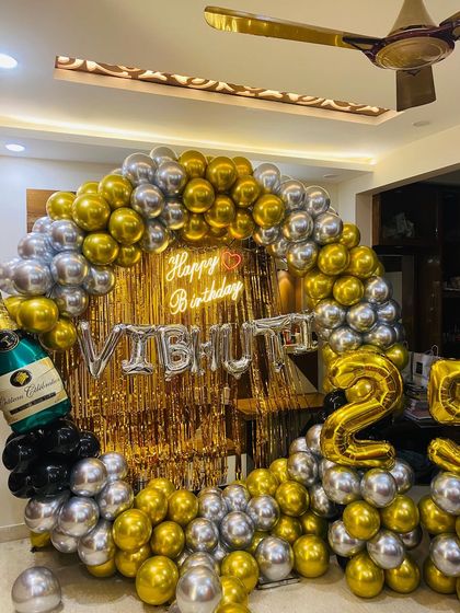 A bold and celebratory 25th birthday setup in a living room. This decor features a massive balloon arch of chrome gold and silver balloons, a gold fringe backdrop, a champagne bottle prop, and foil balloons for the name and age.