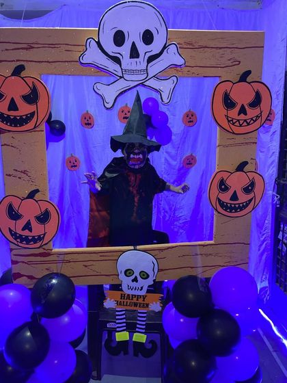 Getting spooky for a Halloween party. This photo booth setup includes a wooden frame, a skull and crossbones cutout, and spooky pumpkin faces, with a child in a witch costume posing inside.