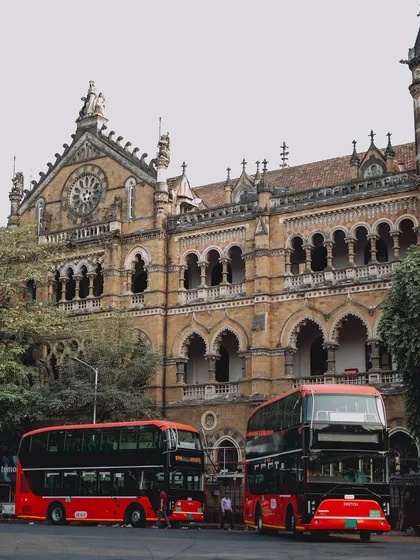 Two of Mumbai's iconic red double-decker buses are parked in front of the magnificent Chhatrapati Shivaji Maharaj Terminus (CSMT). This image captures a blend of the city's modern transport and its historic architecture.