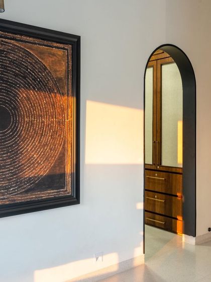 An arched doorway frames a view into the walk-in wardrobe, creating an elegant transition between spaces. The play of golden hour light on the wall highlights the clean lines and the rich texture of the artwork.