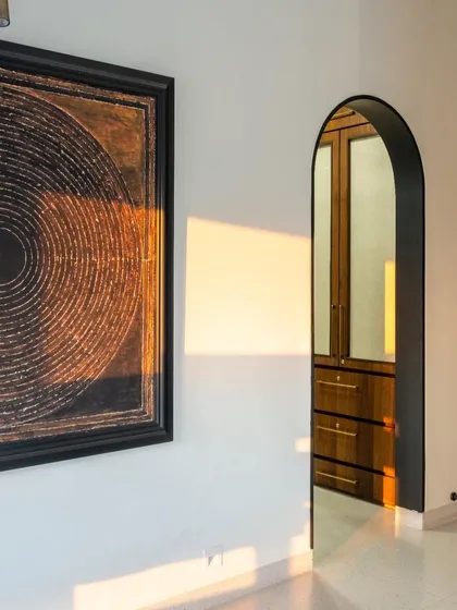 An arched doorway frames a view into the walk-in wardrobe, creating an elegant transition between spaces. The play of golden hour light on the wall highlights the clean lines and the rich texture of the artwork.