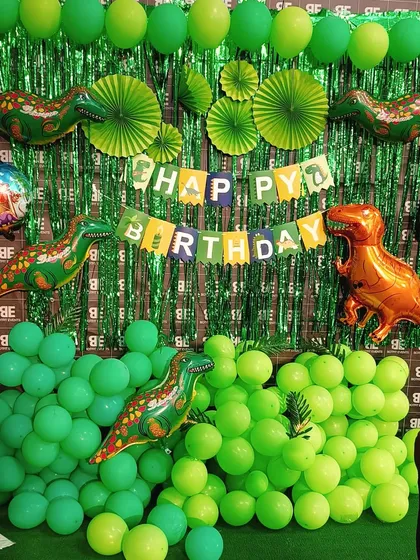 A fun dinosaur themed birthday setup. This decoration features a green shimmer backdrop, various shades of green balloons, and large dinosaur foil balloons to create a prehistoric party atmosphere.