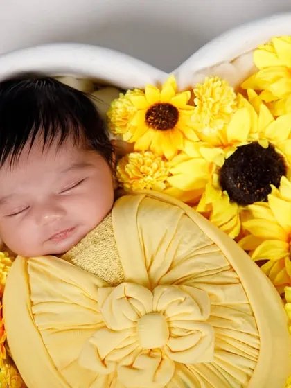 A close-up of a newborn sleeping peacefully in a bed of sunflowers, wrapped in a matching yellow swaddle.