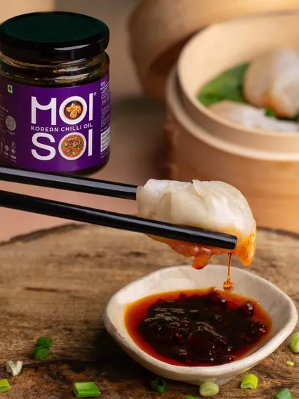 A close-up shot focusing on the action of dipping a momo into Moi Soi's Korean Chilli Oil. This highlights the product in use, creating a craving for both the sauce and the snack.