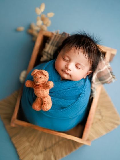 This little boy looks so peaceful in a wooden box, wrapped in a deep blue swaddle and cuddling a tiny teddy bear. The textures create a rustic, cozy feel.