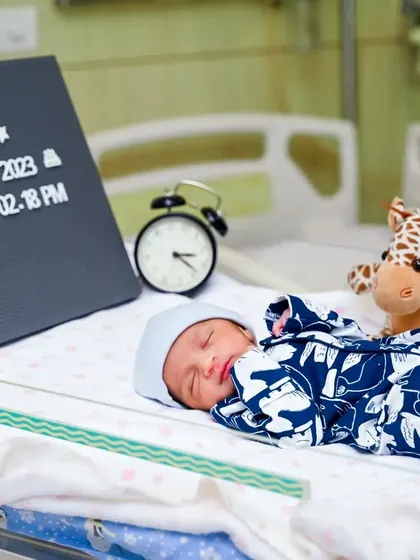 A 'Fresh 48' style hospital photo of a newborn sleeping in a bassinet with a sign announcing his name and birth details.