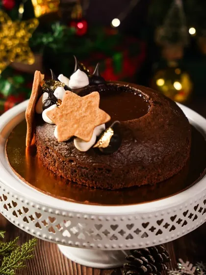 A rich chocolate Christmas cake on a white pedestal, with festive bokeh lights in the background. The dark, moody lighting gives this holiday dessert a luxurious and indulgent feel.