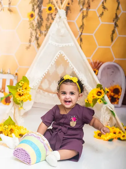 A joyful baby girl in a purple outfit smiles brightly during her sunflower-themed indoor photoshoot.
