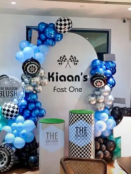 A 'Fast One' racing car theme for a first birthday. This setup includes a custom backdrop, balloon garlands in blue, black, and silver, and props like wheels and checkered flags to bring the race track to life.