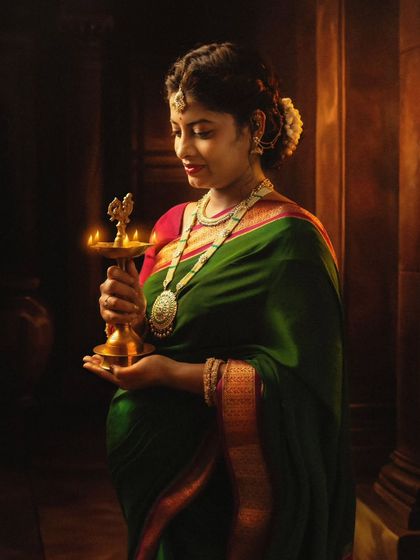 A beautifully lit portrait of a mom-to-be in a traditional green and pink saree, holding a brass lamp (diya) that symbolizes light and life.