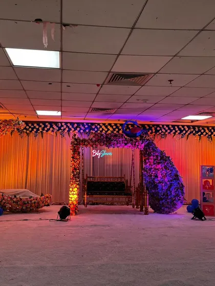 This wide-angle shot shows a complete baby shower stage design in a large hall. The setup includes a floral moon cradle, a decorated jhula swing, and a large balloon backdrop, all arranged on a white carpeted stage with spotlights.