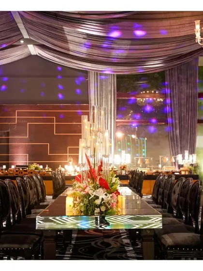 A dramatic, long mirror-top table set for an engagement party, reflecting the purple mood lighting and elaborate floral centerpieces.