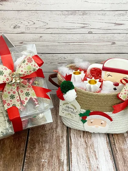 A cozy Christmas hamper featuring a Santa mug and other treats in a woven basket, alongside a box of snowflake-themed gifts.