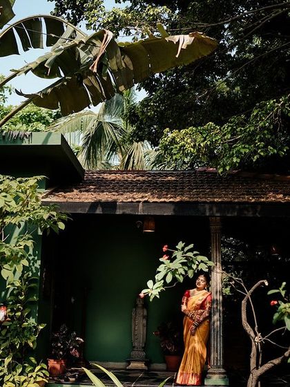 A wide shot of the bride standing on the porch of a heritage building, surrounded by lush greenery.