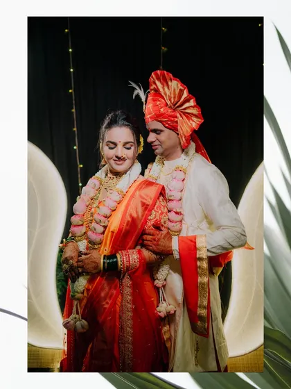 Purva and Dharma framed by elegant decor. The bride's red and orange saree contrasts beautifully with the groom's white sherwani and red pheta.